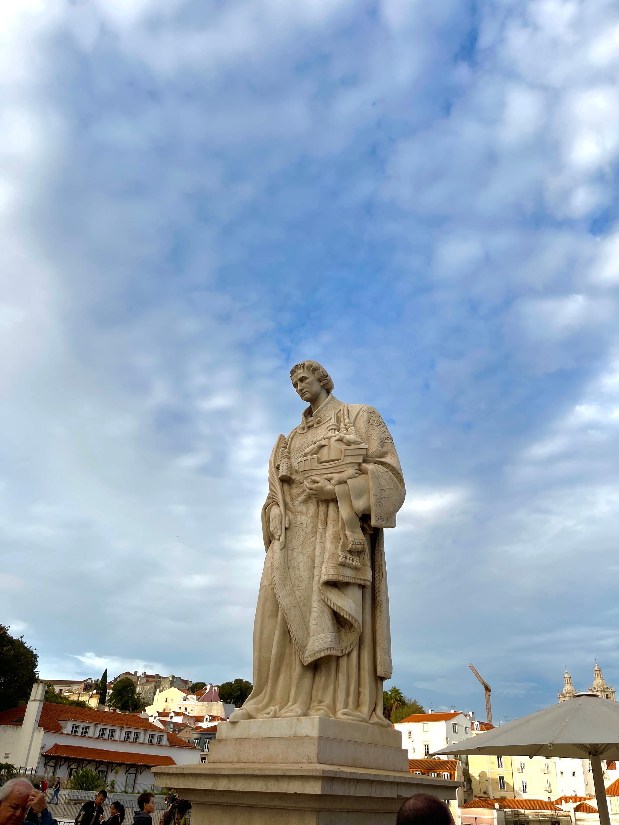 A stone statue of a robed figure stands under a blue sky with clouds, with buildings in the background