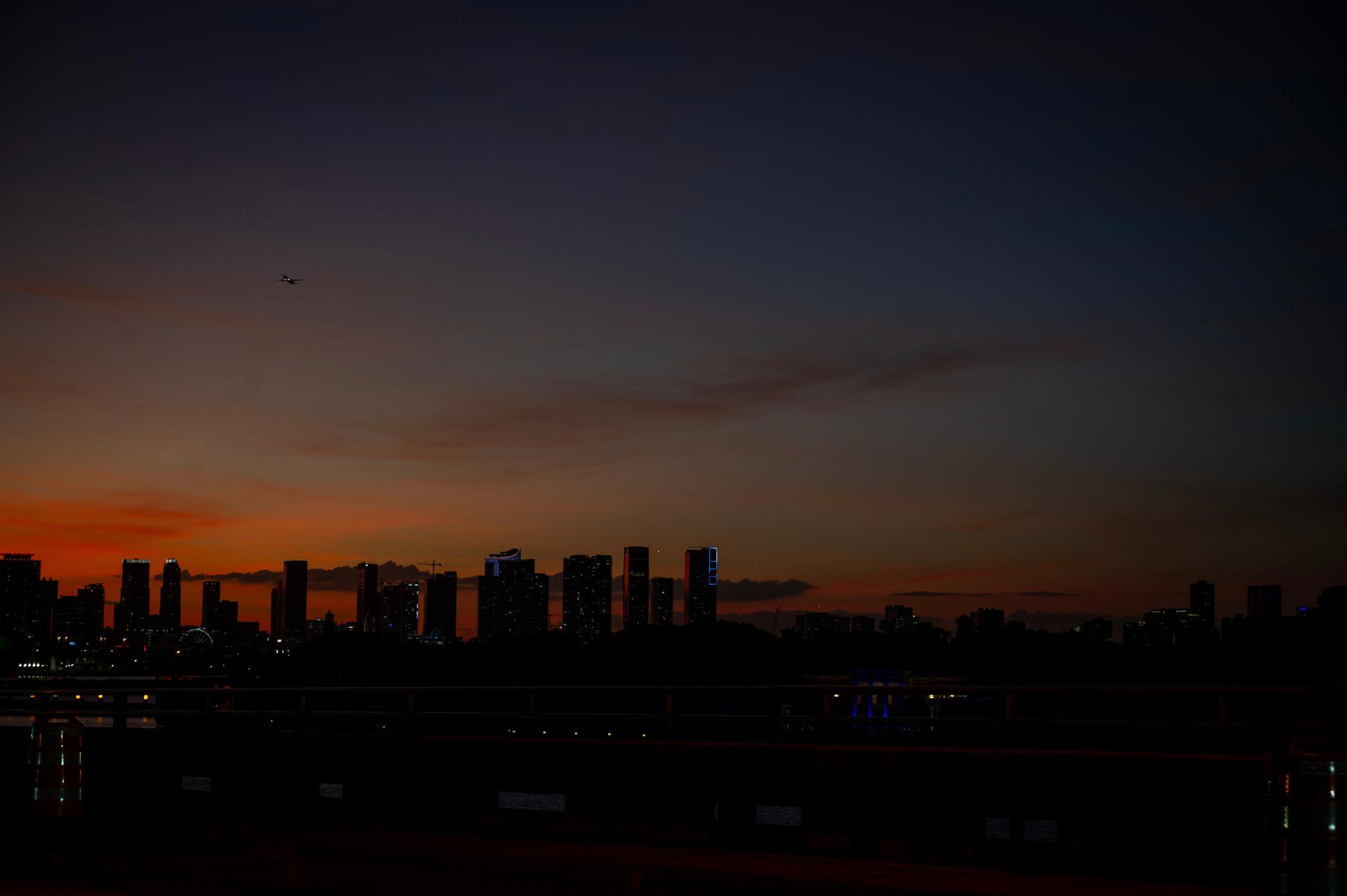 A silhouette of a city skyline against a twilight sky with hues of orange and blue, and a small aircraft visible in the distance