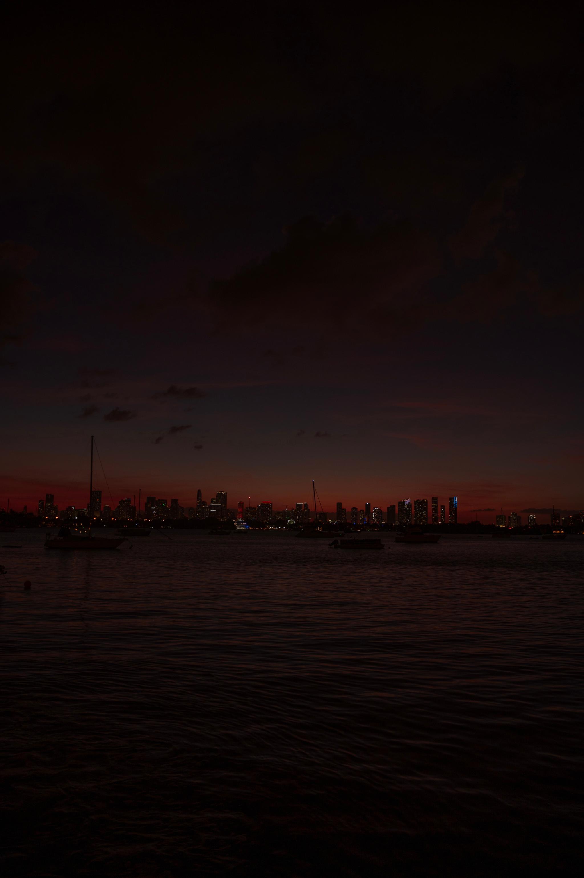 A twilight horizon over a body of water with the silhouettes of buildings and faint lights visible under a dark sky