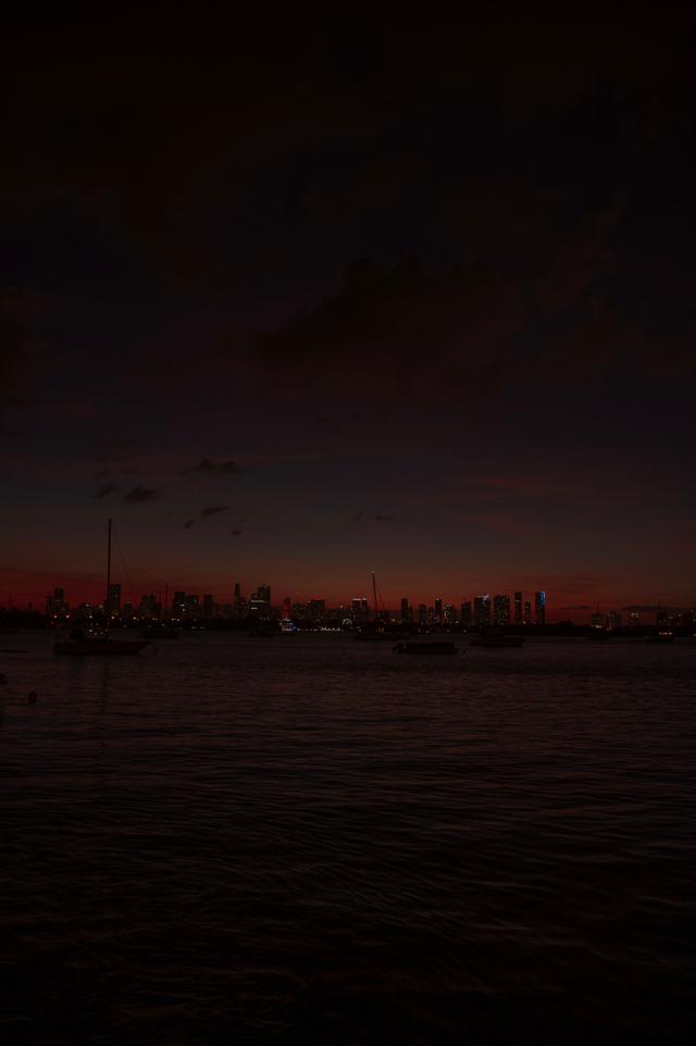 A twilight horizon over a body of water with the silhouettes of buildings and faint lights visible under a dark sky